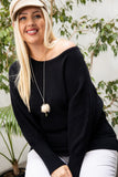 Plus size woman in black off-shoulder sweater, white pants, and beige cap with greenery background