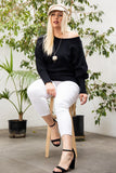 Plus size woman in black off-shoulder sweater, white pants, and black heels, seated outdoors with plants.
